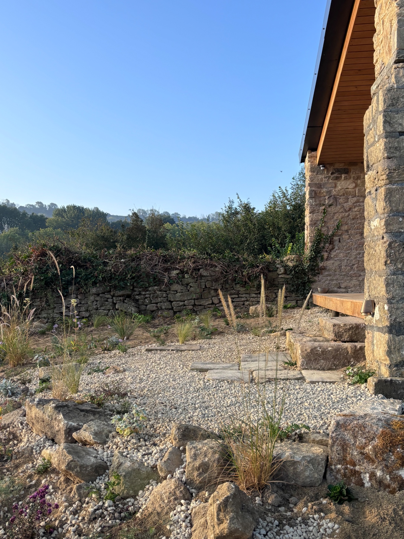 Gravel garden with limestone boulders and ornamental grasses beside a stone studio in low evening sun, landscape design by GHS Spaces, landscape designer in Bristol