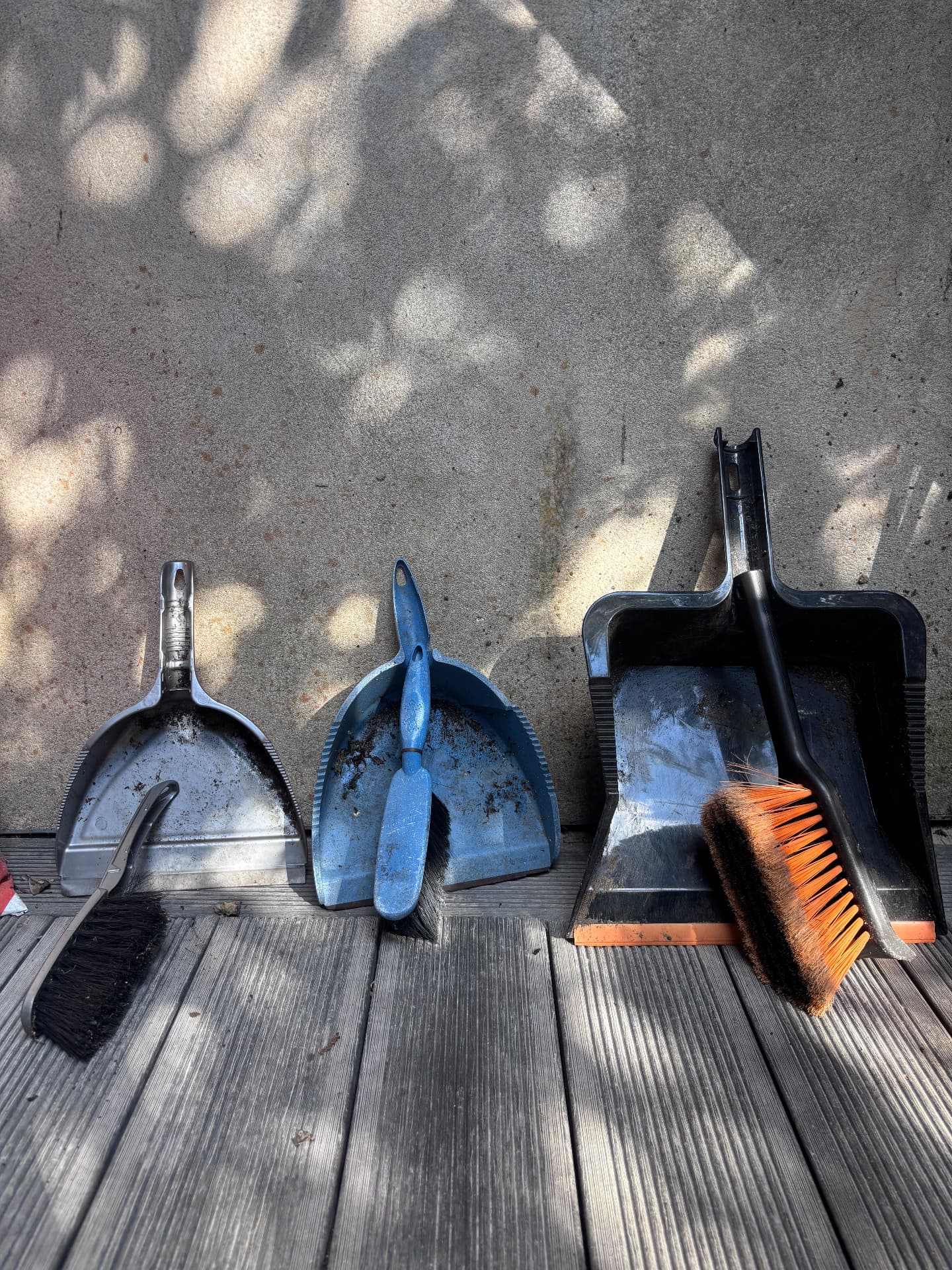 Three dustpans and brushes lined up against a concrete wall in dappled light